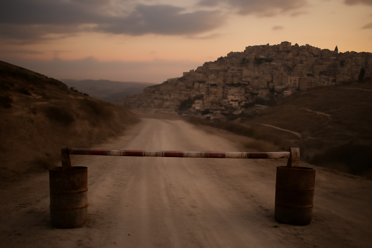 A dusty road leading toward a hillside city in Yemen with a checkpoint barrier in the foreground