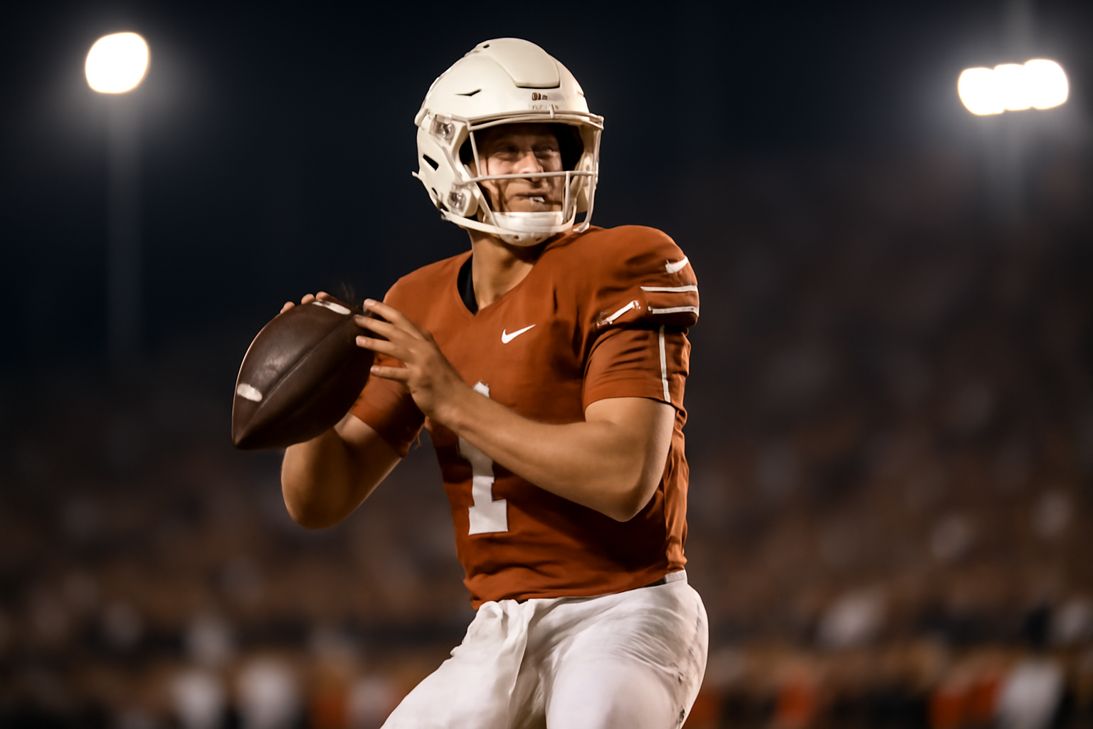 A college football quarterback in a burnt orange jersey drops back to pass under stadium lights during a night game.