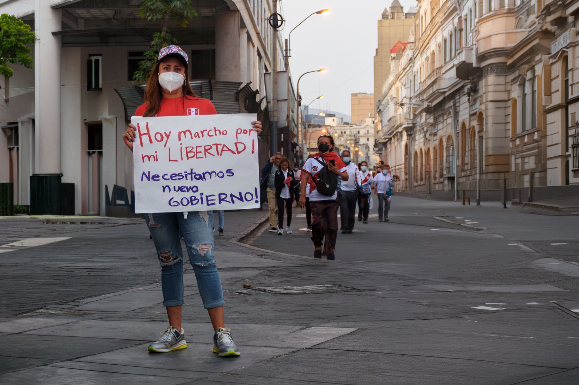 A woman stands outdoors near a Venezuelan flag, looking toward a crowd gathered at dusk