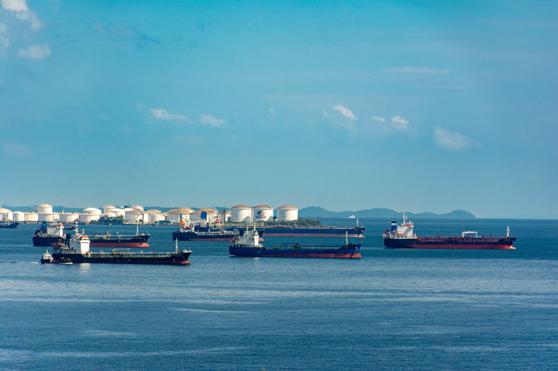 An oil tanker navigating a narrow strait with a refinery visible on the distant shoreline under overcast sky