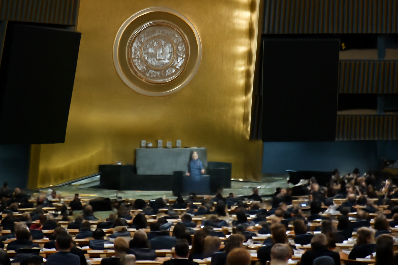 United Nations General Assembly hall with a delegate speaking at the podium beneath the UN emblem