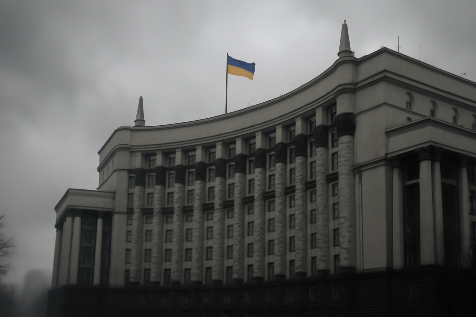 Ukrainian flag and government building under overcast sky with distant smoke