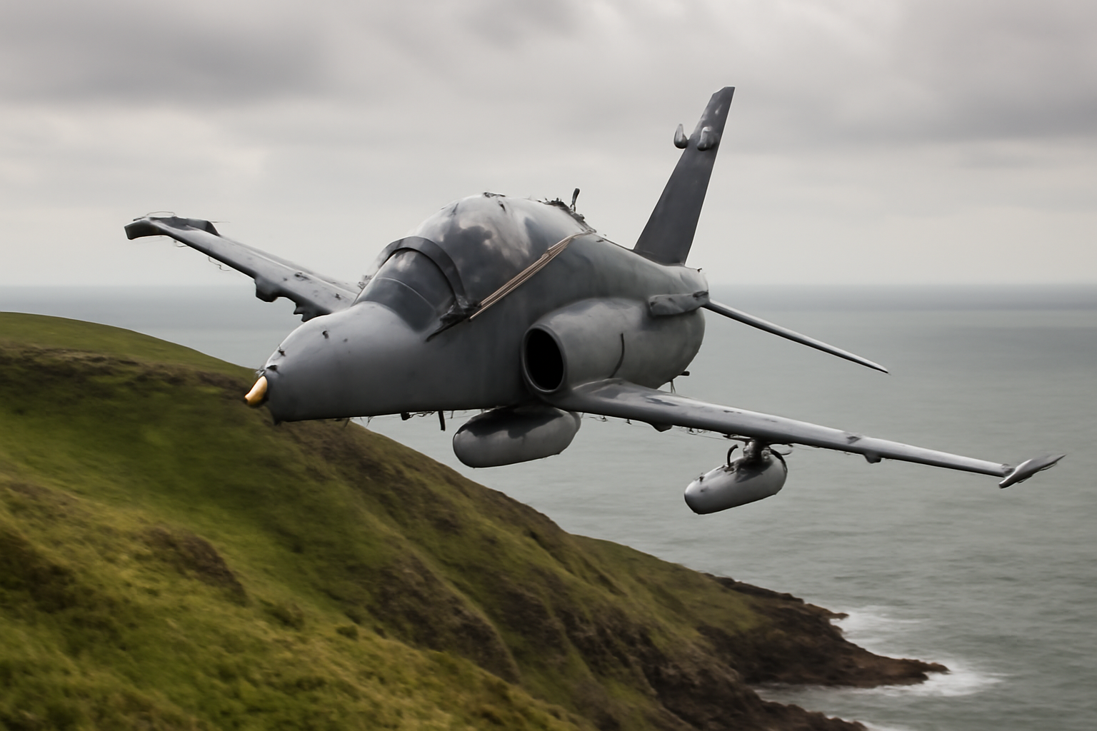 A gray Royal Air Force Hawk T2 twin-seat jet trainer flying low over a rugged Welsh coastline.